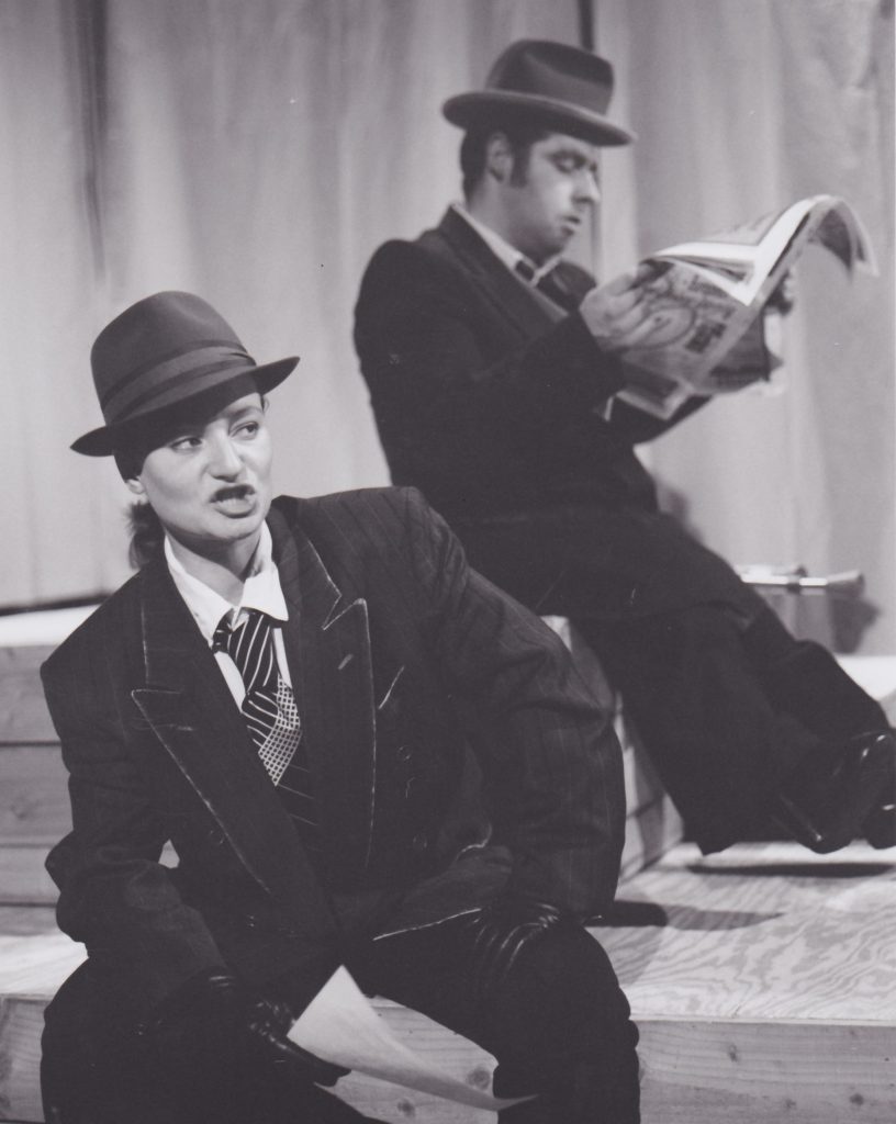 A picture of a woman leaning forward and speaking in a suit and top hat. A man behind her dressed similar reads a newspaper. 