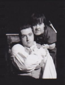 A man sits in a chair looking forwards, a woman stands over his shoulder very close to him. 