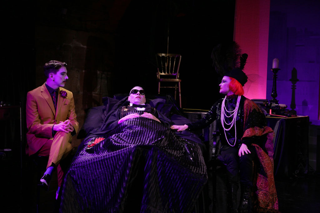 A man in a black suit lies in a bed. Gathered around him are a man in a brown suit with a small moustache and a woman with short red hair dressed in a fancy black dress and a long pearl necklace.