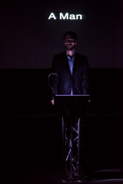 A man in a suit stands at a lecture-like podium. Behind him we see a projection that reads 'A Man'. The stage is lit very dark so most of the man's features are obscured.