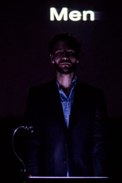 A shadowy close up of a man in a suit at a podium. The word 'Men' is projected behind him.