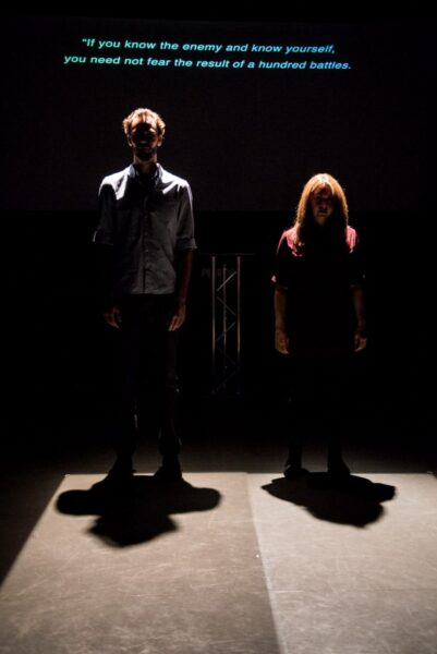 A man and a woman stand on stage. They are being lit from above, casting shadows that make their faces difficult to see. A projection behind them reads: 'If you know the enemy and know yourself , you need not fear the result of a hundred battles.'