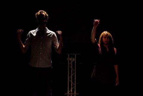 A man and woman stand on a dimly lit stage. They have their arms in the air as if in celebration.