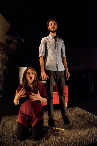 A tall man stands on a grey rug looking off into the distance. He is wearing grey trousers and a blue shirt. He has brown hair and a short brown beard. A woman is on her knees beside him, she appears to be signing. She has long brown hair and is wearing a purple dress.