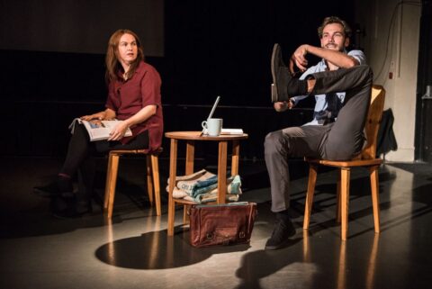 A man and a woman sit on wooden chairs. They are both white and appear to be in their 30s. There is a small wooden table in between them with a laptop and a mug on it. The woman has a magazine on her lap and is looking at the man who has one of his legs halfway in the air as he unlaces one of his shoes