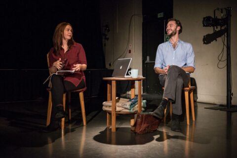 A man and a woman sit on wooden chairs. They are both white and appear to be in their 30s. There is a small wooden table in between them with a laptop and a mug on it. The man is laughing while the woman looks over at him amusedly.