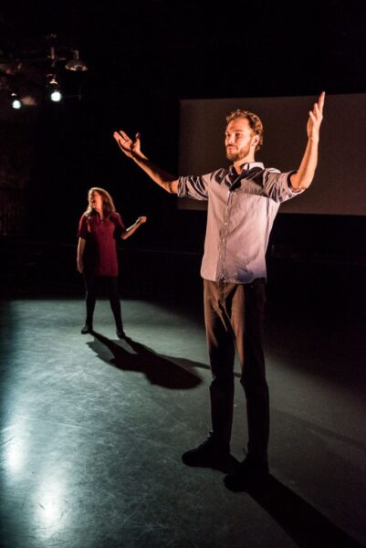 A tall white young man stands on a bare stage with his arms in the air and light shining down on him. There is a woman in the background, she appears to be in the middle of talking.