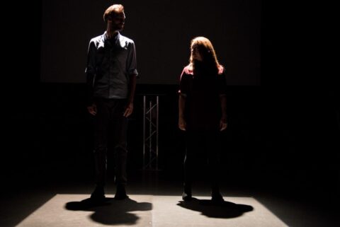 A man and woman stand beside each other on a bare, dark stage. They are looking at each other