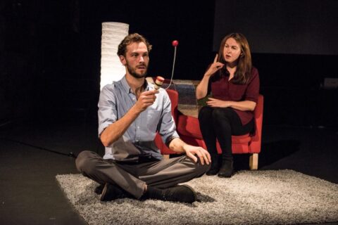 A man sits cross legged on grey shag run. He is playing with a cup that has a ball on the end of a string attached to it. A woman in a red armchair sits near him and appears to be signing. The man appears to be in his early 30s, he has brown hair and a short brown beard. He is wearing grey trousers and a blue shirt. The woman also appears to be in her 30s, has long brown hair and is wearing a purple dress with black tights.