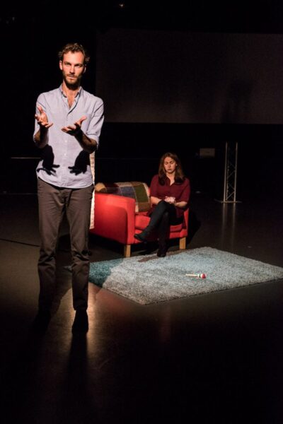 A tall young man stands in the foreground with his hands out. A woman in a purple dress sits in the red armchair with her eyes cast down.