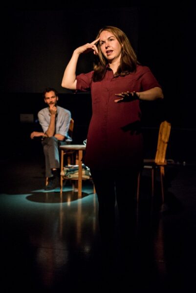 A woman stands signing at the front of the stage. She is holding out one hand while pointing the other to her head. She appears to be in her 30s, white, has long brown hair and is wearing a purple dress with black tights .A man is sitting on a wooden chair in the background. He is leaning forwards and appears to be listening intently.