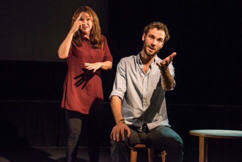 A young white man sits on a wooden chair. He has one hand on his knee and is holding the other out to the audience. A woman stands beside him, signing. She appears to be in her 30s, white, has long brown hair and is wearing a purple dress with black tights.