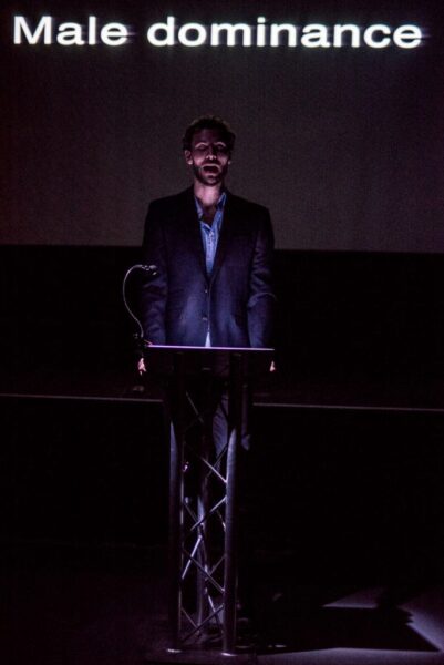 A man in a suit stands at a darkly lit lecture podium. The words: 'male dominance' projected on the screen behind him.