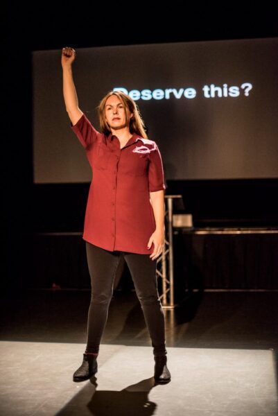 The woman stands at the front of the stage with one of her fists in the air. Behind her the words 'Deserve this?' are projected. She appears to be in her 30s, white, has long brown hair and is wearing a purple dress with black tights.