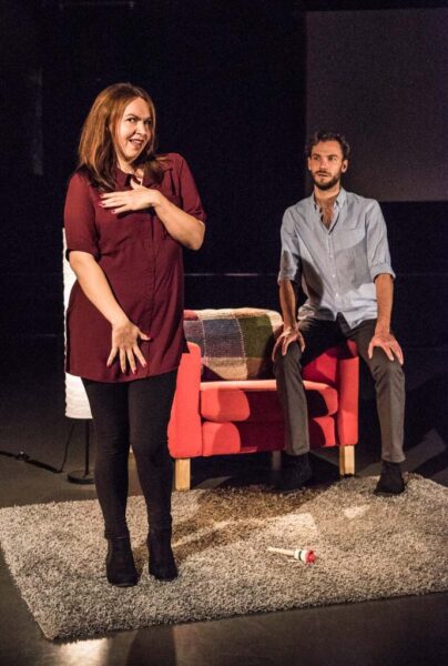 A woman is standing on a grey rug. She is suggestive running one of her hands across her chest and the other down her thighs while a man sits on the arm of a red armchair and watches.