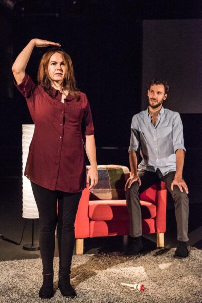 A woman is standing on a grey rug. She appears to be signing, she is standing up very straight and holds one hand out flat over her head. A young man sits on the arm of a red armchair and watches with admiration.