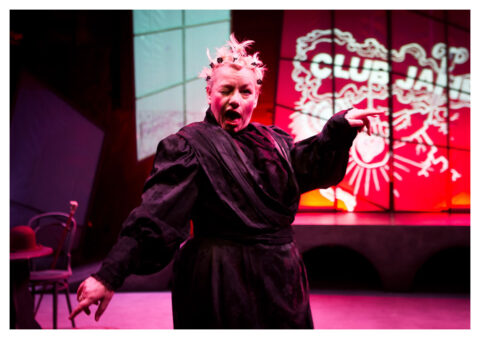 A middle aged white woman in a black dressing gown strikes a pose on stage and gives an exaggerated wink. She has short bleach blonde hair, that has been twisted into multiple bunches held in place with black hair clips. Behind her there is a bright orange and white projection that reads 'Club Jane' . Across from this there is a small wooden chair with a table next to it. The chair has a cane resting against it and the table has a purple hat on it.