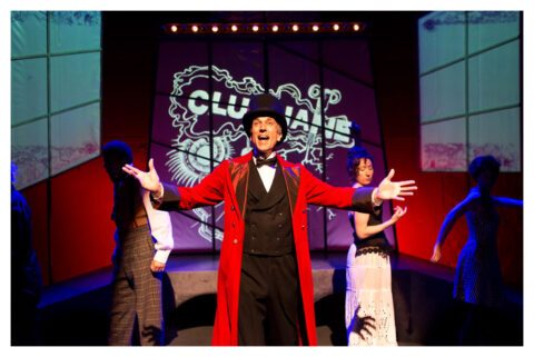 A man in a long red coat and a black top hat stands at the front of the stage with his arms out. There are actors in the background wearing what looks like Victorian style clothing.