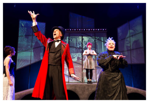 A man in a long red coat and a black top hat extends one arm up to the sky, he appears to be singing. A woman in a Victorian style black dress stands beside him signing. Behind them there is a sprawl of projection screens showing different images.