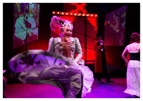 A middle aged white woman in a big poofy violet dress and a big red feather in her short blonde hair is dancing.