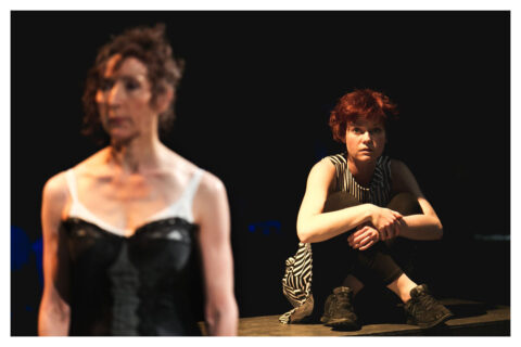 A young white actress with short red hair sits on the floor with her knees brought up to her chest. She looks on at another actress in the foreground. This actress appears to be older with curly dark hair that is tied back. She is wearing a white dress with a large black corset.