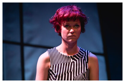 A close up of an actress in a black and white striped dress. She looks angrily above her. She is white, appears to be in her late 20s and has short deep red hair with a fringe.