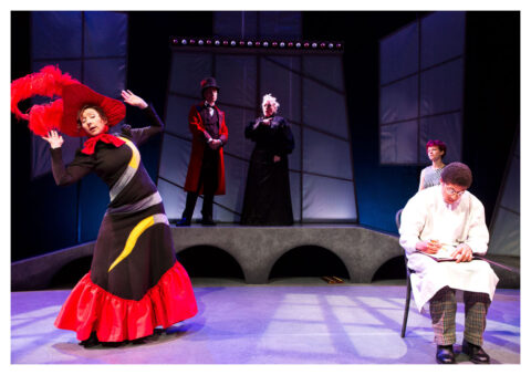 An actor in a long white tunic and glasses sits at the front of the stage writing. A woman to his right is wearing a black dress that has a large red skirt and a large red feathered hat.and stands with her arms up.