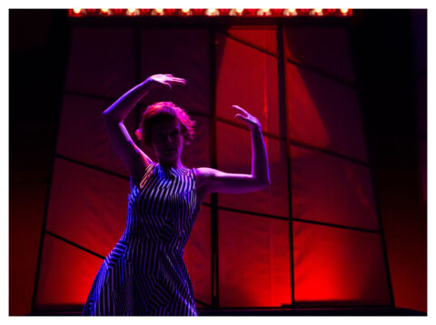 The stage is lit a dim red. An actress in a black and white striped dress stands with her arms in the air.
