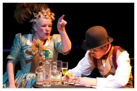 A man and a woman sit at a table filled with half empty bottles and glass. The man is looking down as he appears to be writing and the woman is pointing at someone out of frame in an accusatory manner. The woman is white, middle aged and has short blonde hair. She is wearing a poofy Victorian style light blue dress that has a large yellow flower at the front of it. She also has large brown feathers attached to the back of her head. The man's face is largely obscured as he is looking down, but he appears to be young. He is wearing a brown hat with a white shirt and a brown and red waistcoat.
