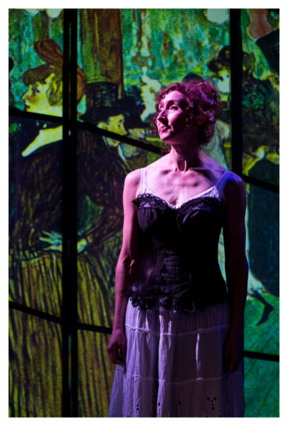 A woman in a black corset and a long white skirt stands in front of a stained glass window. She is white, appears to be middle aged and has curly brown hair that is tied up.