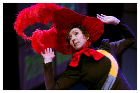 A woman in a black dress and a large red feathered head leans back with her arms up.