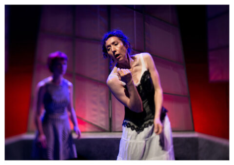 An actress in a black corset and a long white skirt appears to be singing at the front of the stage. She is white, appears to be middle aged and has curly brown hair that is tied up.