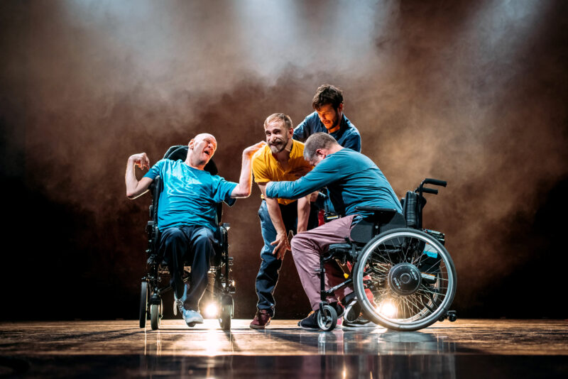 Four male performers clustered on a slightly smokey looking stage. Two of them are in wheelchairs, the other two are standing. Their ages appear to range from young to middle aged and they are dressed in various coloured shirts and jeans. They all appear to be reaching out to grab a man in a yellow T shirt who is standing in the middle of them all.