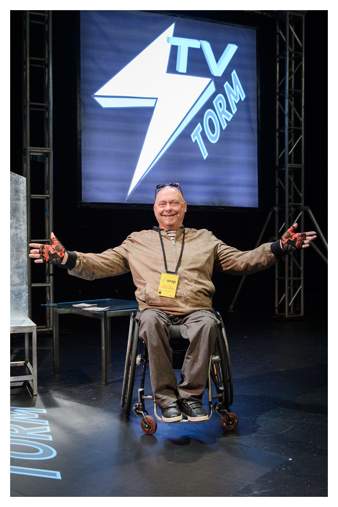 A photo of former artistic director Garry Robson on stage. He is posing with his arms out. He is a white man in a wheelchair, he is bald and appears to be in his early 50s.