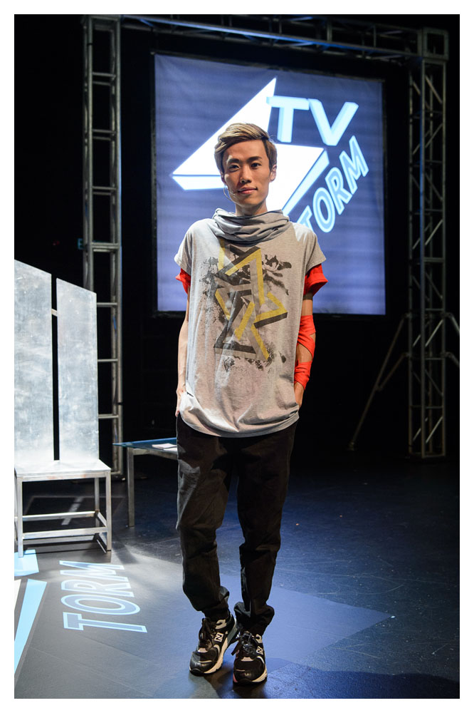A close up of an actor in baggy grey and black clothes. He has lots of orange bands wrapped around his his left arm. He appears to be Asian, in his late 20s and he has brown hair with a long side fringe.