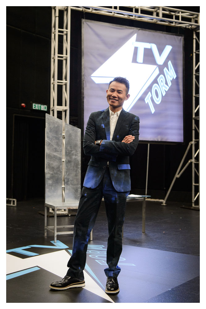 A close up of an man on set in a smart blue suit. He is Asian, appears to be in his mid 30s and has black spiked up hair.