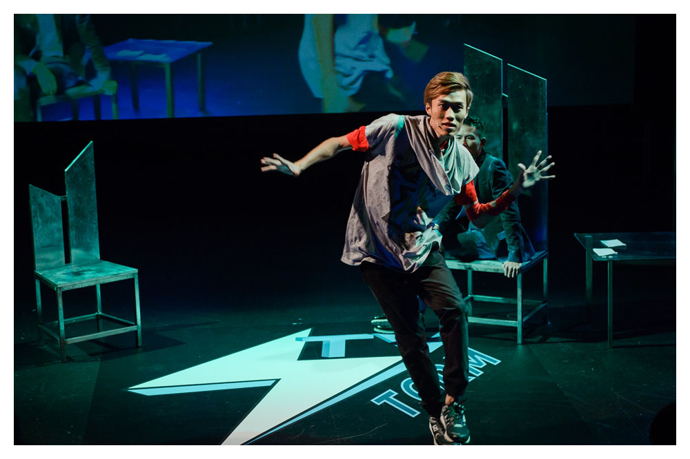 A green wash covers the stage as an actor in raggy clothing dances. He appears to be Asian, in his late 20s and he has brown hair with a long side fringe. A geometric logo is projected on the stage floor underneath him. Behind him there are some scattered wooden chairs. An actor sits in one of them and looks on. He is Asian, appears to be in his 30s, wears a blue suit and has black spiked up hair.