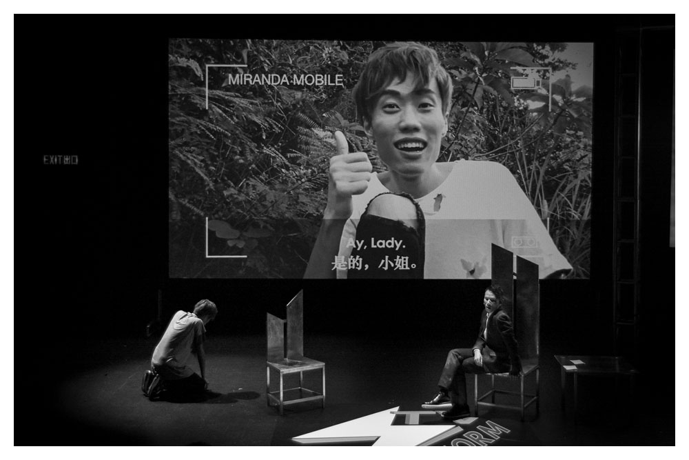 A black and white photo where two actors are on stage. One is on a shoddily made driftwood chair and the other is crouched on the floor. There is a projection on the back of the stage of a man being interviewed on a camera phone. He is giving a thumbs up.