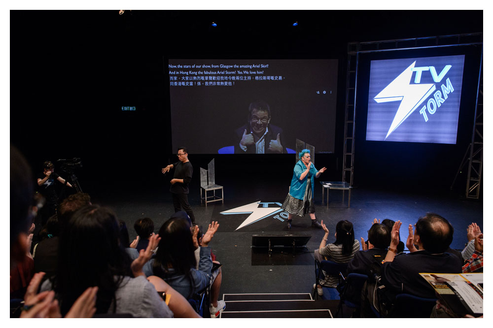 A photo taken from in between rows of audience members, they all appear to be in the middle of clapping. On stage, there are two people, one white man and a white woman who appear to be in the middle of signing. On stage there are a few high backed chairs, a logo that says 'Tv Torm' on both the floor and on a screen suspended at the back. In the middle of the stage there is a large projection screen, showing a man in a suit and glasses giving two thumbs up.