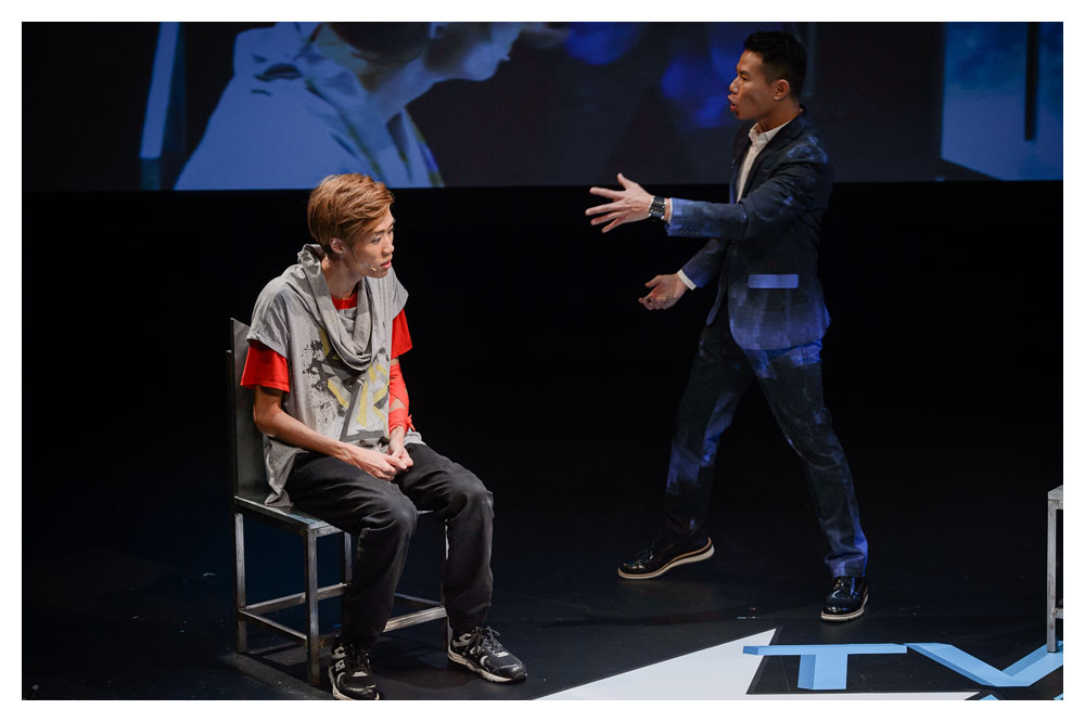 A young Asian actor sits on a chair on stage staring into the distance. A slightly older actor in a suit stands next to him and appears to be talking.