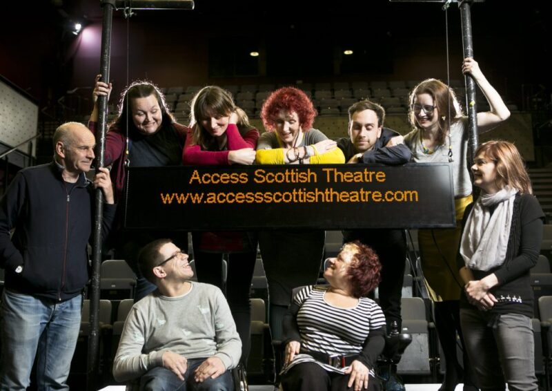 A group of people gathered on a stage with the seating bank behind them. They are gathered around a captioning unit that says 'Access Scottish Theatre. www.accessscottishtheatre.com
