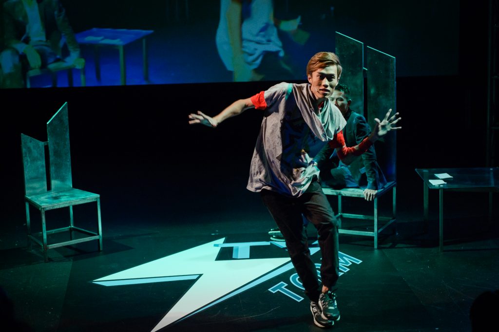 A green wash covers the stage as an actor in baggy ragid clothing dances. He appears to be Asian, in his late 20s and he has brown hair with a long side fringe.