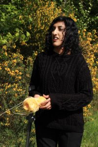 A photo of Aminder Virdee that looks to have been taken in a garden. She is an Asian woman who appears to be in her early 20s. She is wearing a black jumper and has long, wavy black hair. 