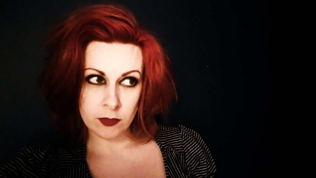 Profile of Jen, she has red hair in a short bob cut with thick make up around her eyes with which she looks out the corner of here eyes into the distance.