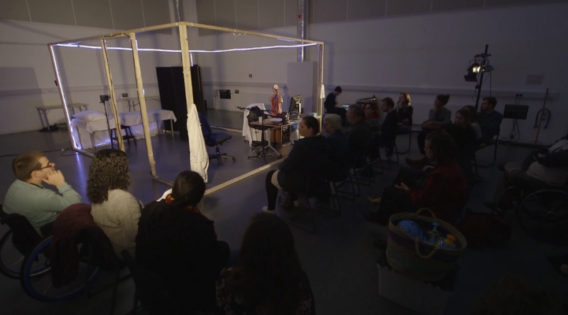 The image shows the frame of a large wooden cube, with no walls so we can see inside. It has been made to look like a doctors office; there is a desk with computer and piles of files; a medical model of the human body; a medical looking bed separated from the desk by a screen. On the other side of the image an audience sits a watches.