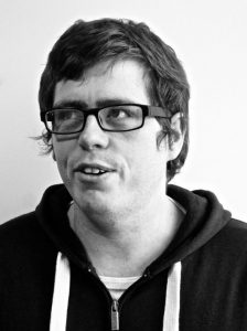 A black and white photo of Stuart Pyper. He appears to be a white man in his 30s. He has short brown hair and thick rectangular glasses and is wearing a hoodie.