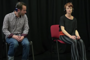 A man and a woman sit in front of a black curtain, they are both wearing large black headphones. The man appears to be white, in his 30s with black hair and stubble, he is looking over at the woman. The woman is also white, appears to be in her 30s and has short brown hair. She appears to be lost in whatever she is listening to.