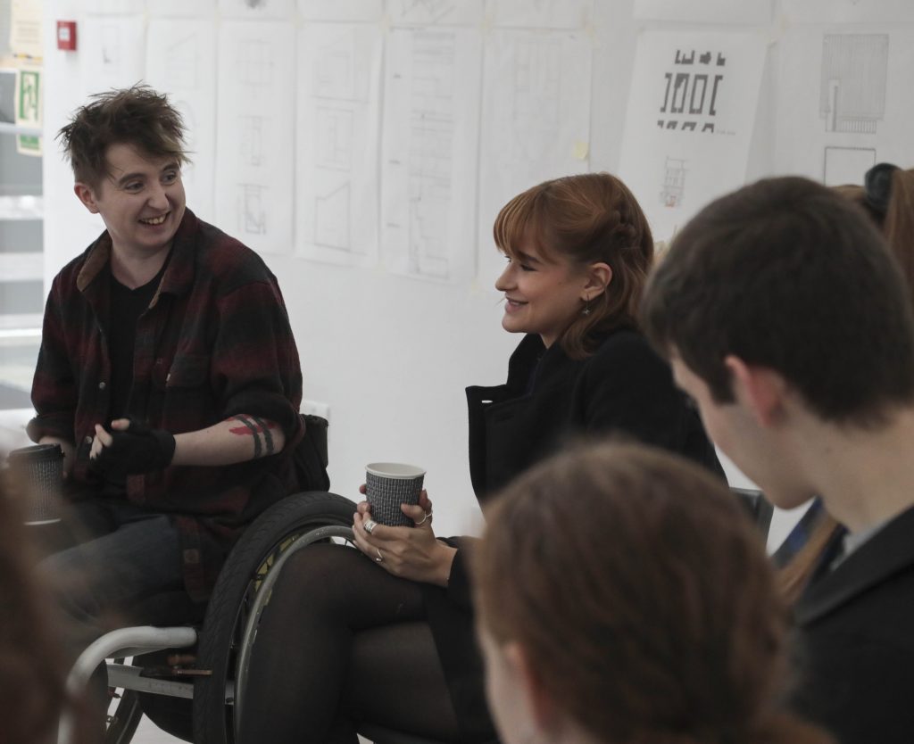 A shot of people in a workshop. We see a young man on the left, he is wearing a checked red and black shirt. He is smiling at a woman next to him, she has brown hair and is holding a cup of tea. In the background we see a white wall covered in various pieces of paper with plans on them.