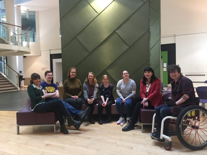 8 young people sit on a large sofa together and smile for a photo.