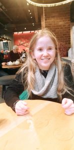 A photo of Sorcha Pringle. She is sitting at a table and is wearing a black long sleeve top. She has long blonde hair and is smiling at the camera.